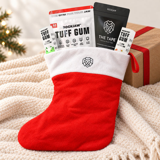 The ROCKJAW Stocking, a red felt Christmas stocking with a white cuff and the ROCKJAW lion logo, leaning against a kraft-wrapped present on a cream knitted throw, filled with a Tuff Gum Strawberry Smash 60 piece pouch, The Tape mouth tape and two small Tuff Gum packs.