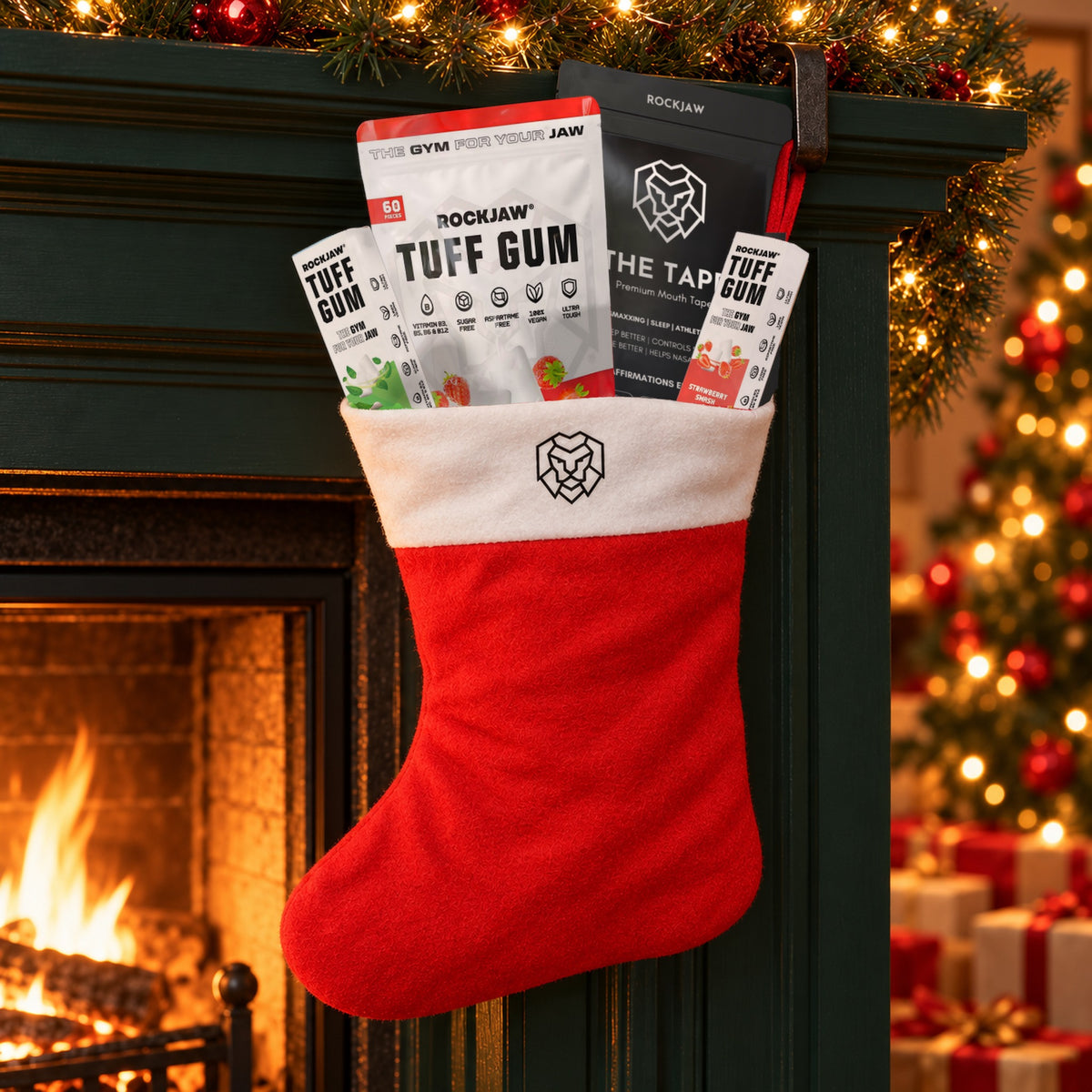 The ROCKJAW Stocking hanging from an iron hook on a dark green fireplace mantel at night, with Tuff Gum Strawberry Smash, The Tape and two small Tuff Gum packs tucked into the cuff, a fire glowing beside it and a lit Christmas tree behind.