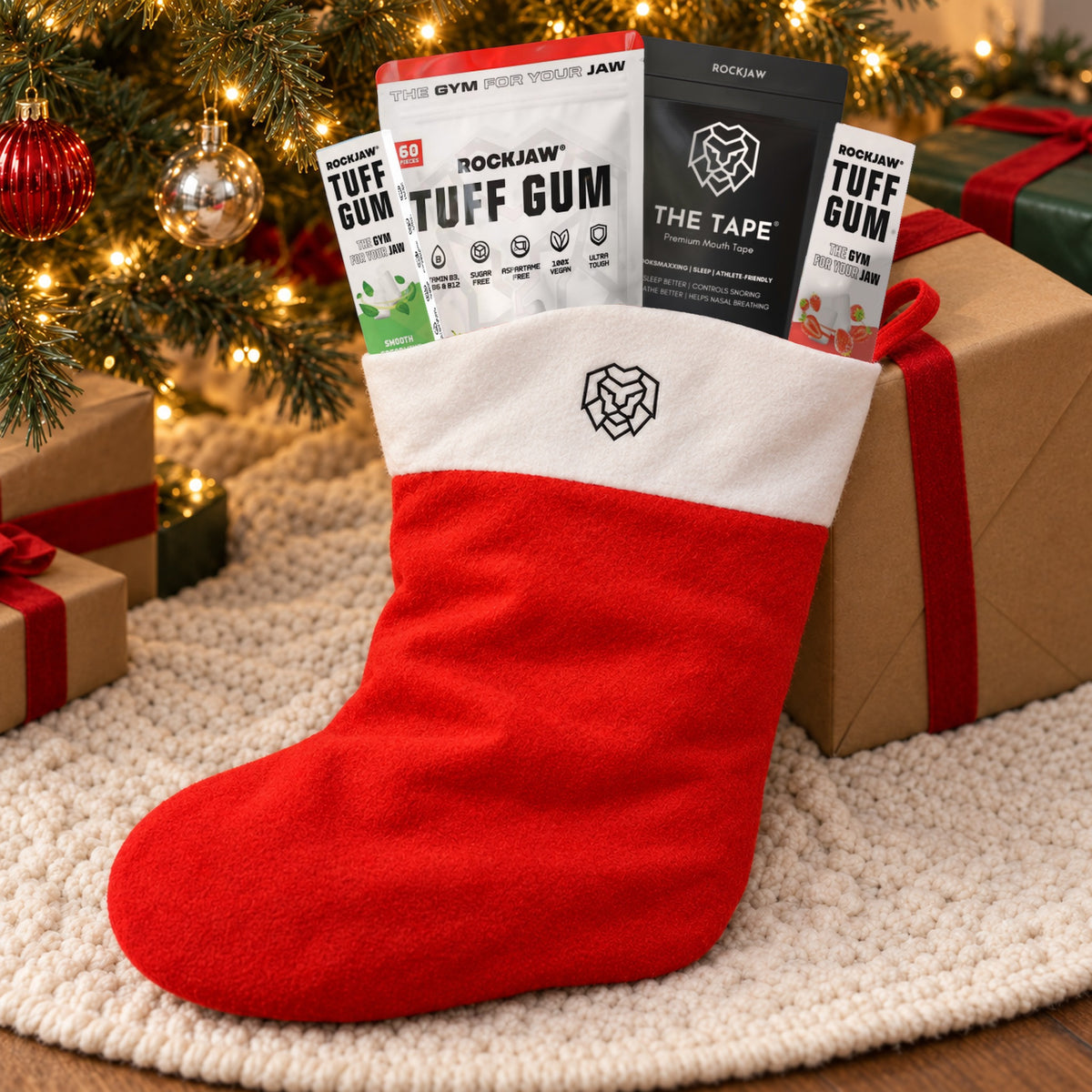 The filled ROCKJAW Stocking resting on a cream knitted tree skirt against a kraft-wrapped present under a Christmas tree, with Tuff Gum Strawberry Smash, The Tape and two small Tuff Gum packs tucked into the cuff.