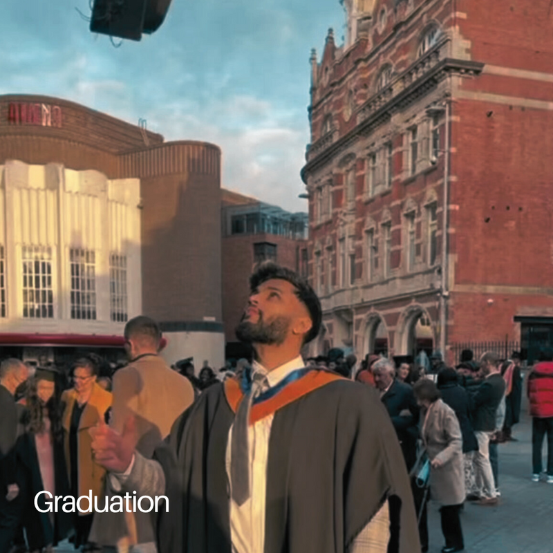 Sach at graduation in gown and hood, outside a red-brick building